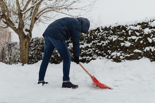 Man clearing snow with a red shovel in a winter backyard setting.